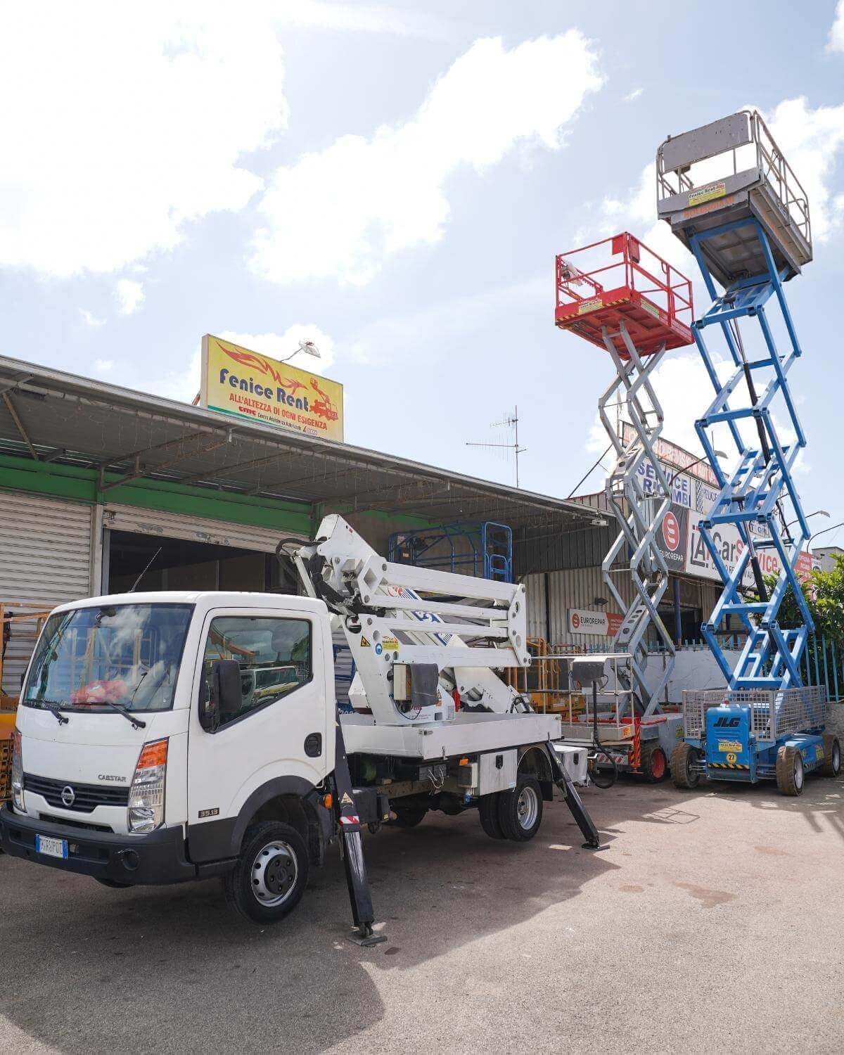 Camion bianco con piattaforma aerea, due piattaforme a forbice e la facciata di un edificio.