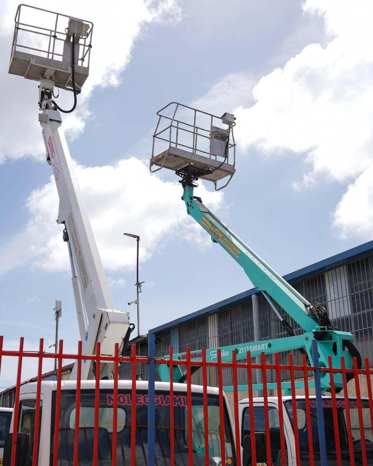 Due carrelli elevatori con piattaforme rialzate dietro una recinzione rossa, contro un cielo nuvoloso.