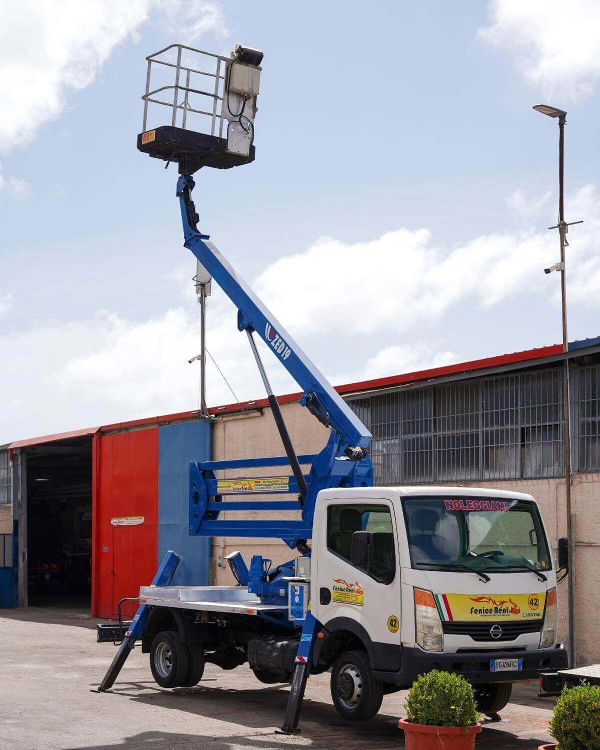 Piattaforma aerea montata su camion con braccio blu, cestello bianco, gambe di supporto estese, vicino a un edificio in una giornata di sole.