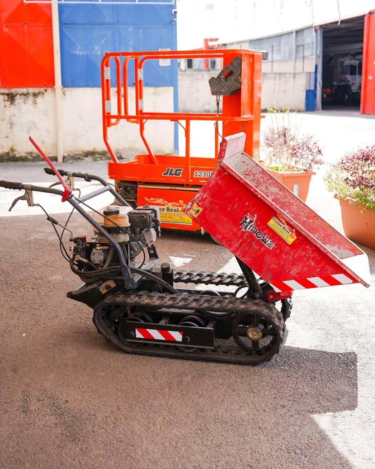 Dumper cingolato rosso con cassone rialzato; sollevatore arancione sullo sfondo. Esterno.