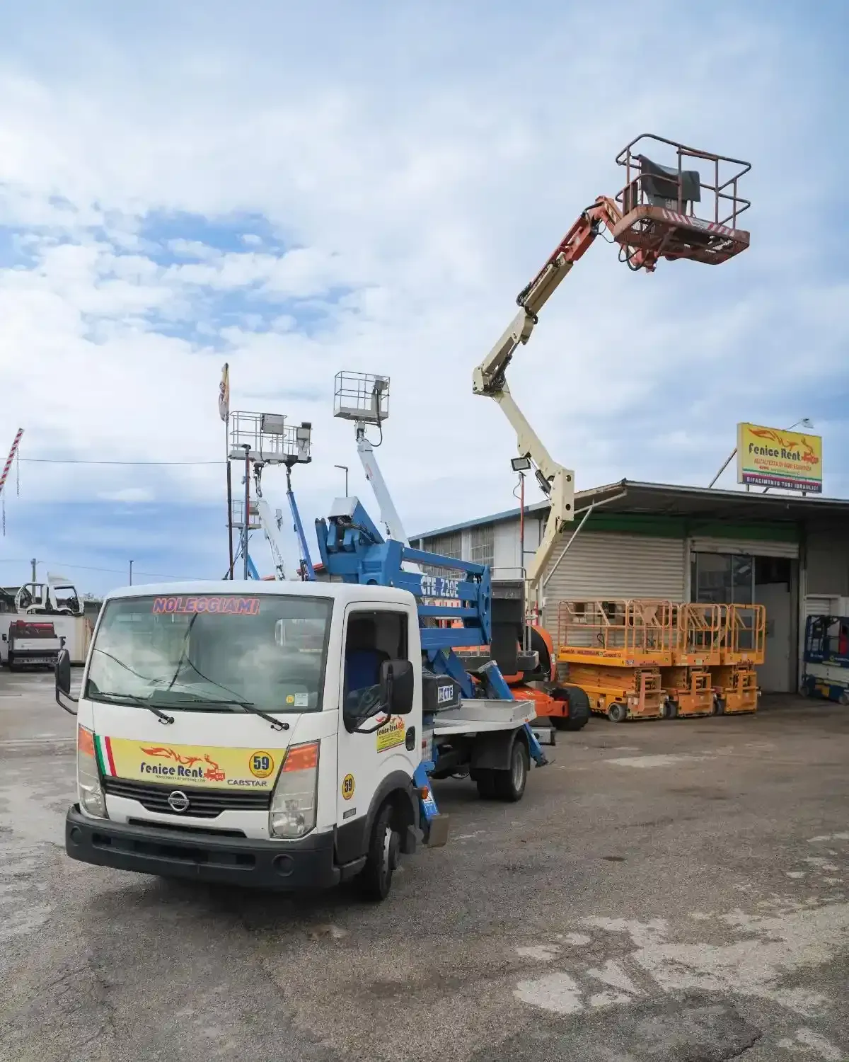 Camion bianco con piattaforma elevatrice blu estesa; parcheggiato di fronte a un edificio con attrezzature. Giornata nuvolosa.