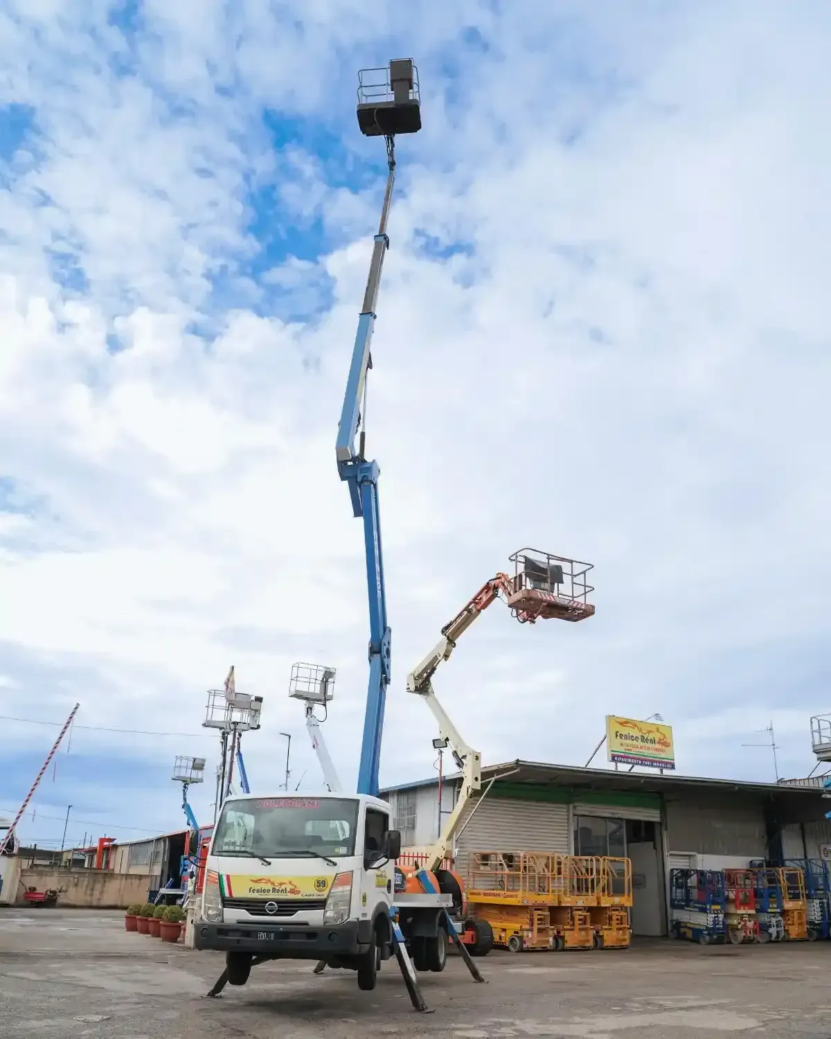 Piattaforma aerea montata su camion con braccio esteso blu e bianco, accanto a una piattaforma più piccola arancione e bianca. Esterno, cielo nuvoloso.
