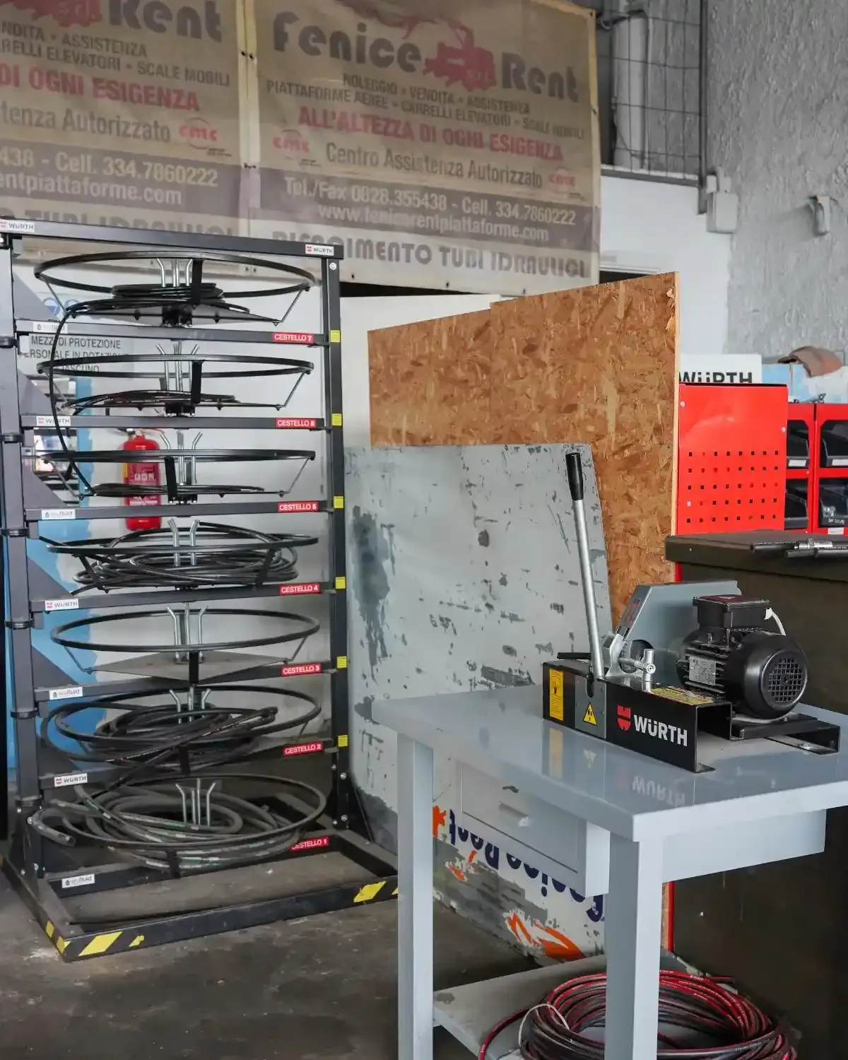 Scaffale metallico con fili arrotolati accanto a un tavolo da lavoro con una macchina. Ambiente da officina.