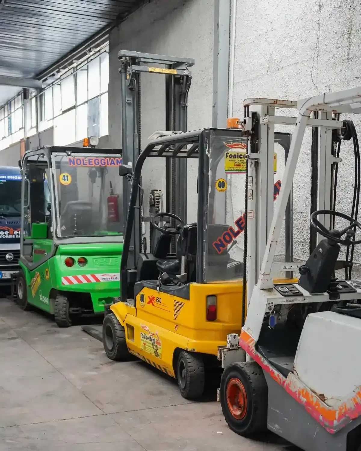 Tre carrelli elevatori parcheggiati in un magazzino: verde, giallo e bianco, visti di lato.