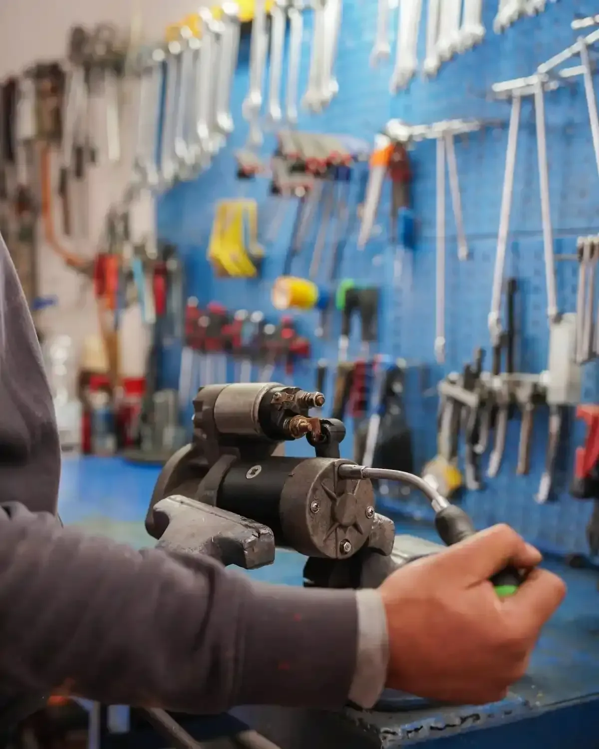 Meccanico che tiene in mano un motorino di avviamento in un'officina con attrezzi su una parete blu.