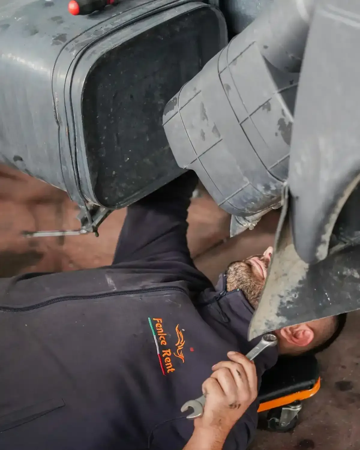 Meccanico al lavoro sotto un veicolo, con in mano una chiave inglese. Indossa un'uniforme scura con logo, in un'officina.