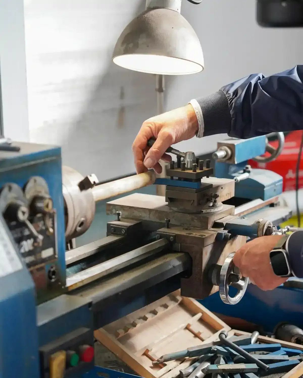 Persona che aziona un tornio, con in mano un pezzo di legno. La macchina è blu, con una luce intensa dall'alto. Ambiente di officina.