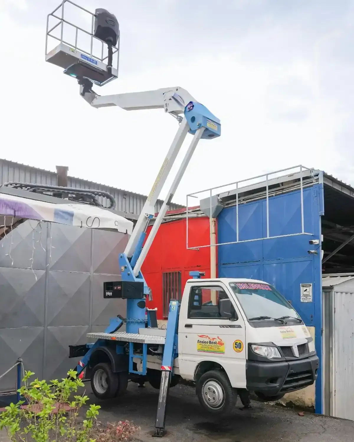 Camion bianco con piattaforma aerea sollevata. Parcheggiato accanto a un muro e a un edificio blu.