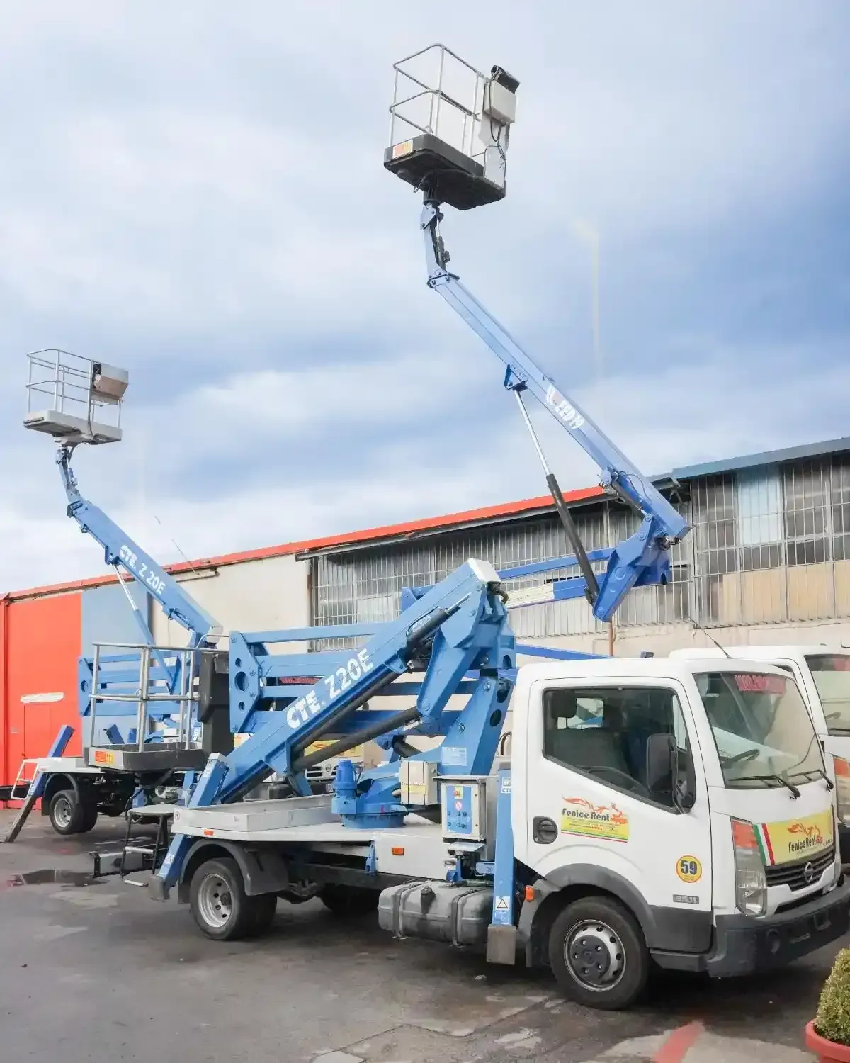 Autocarro elevatore blu con piattaforma rialzata, parcheggiato vicino a un edificio in una giornata nuvolosa.