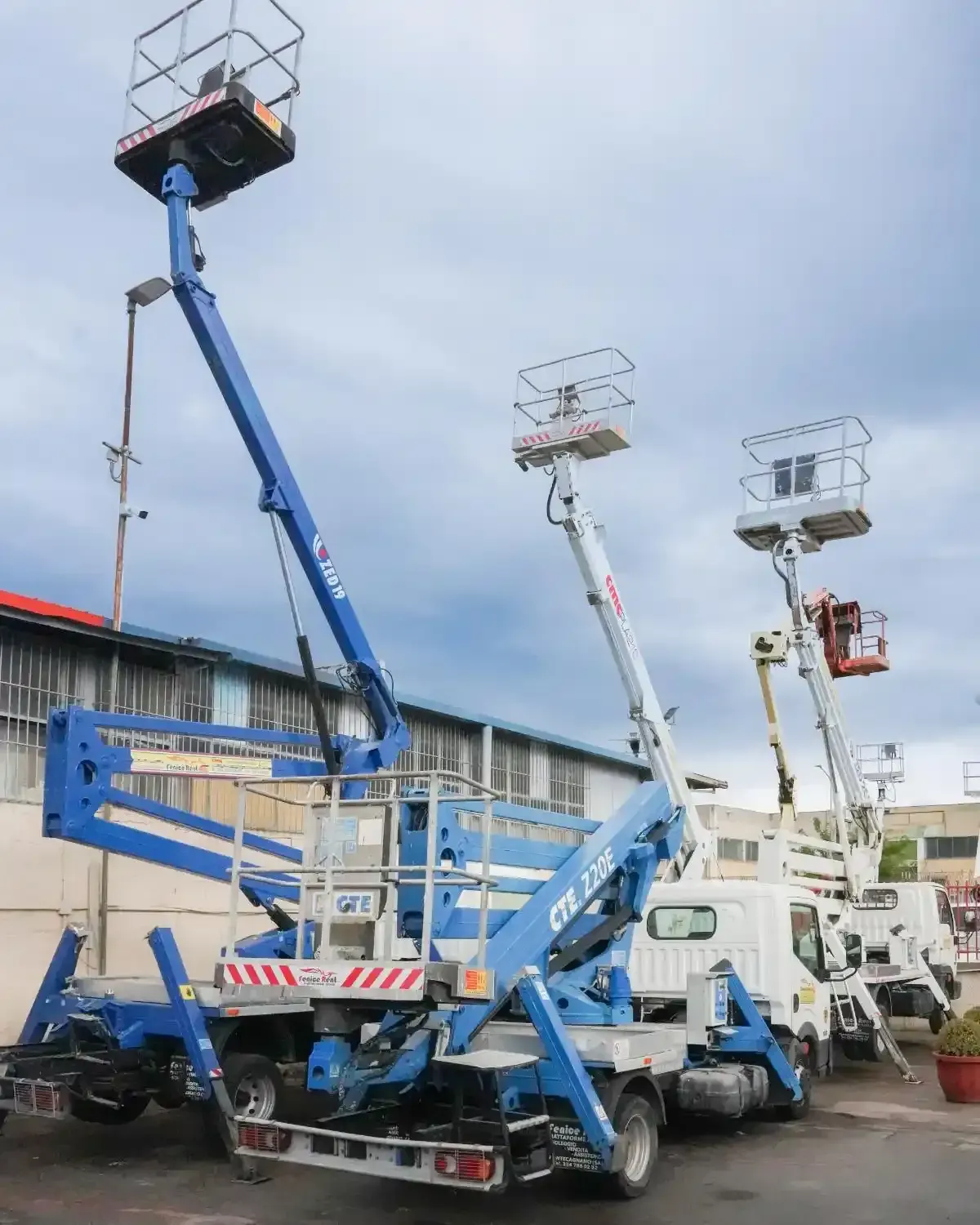 Tre piattaforme aeree montate su camion parcheggiate all'aperto. Piattaforme blu, bianche e rosse si estendono verso l'alto contro un cielo nuvoloso.