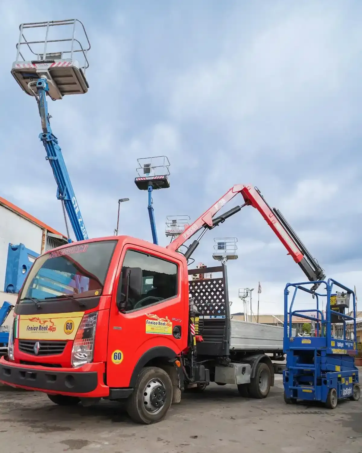 Camion rosso con gru e piattaforme sollevate, un'altra piattaforma e una piattaforma aerea a forbice blu sollevata in un cortile.