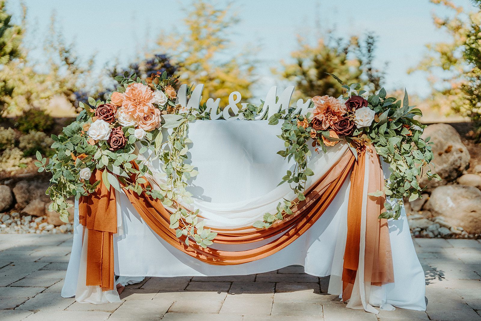 Wedding sweetheart table, white tablecloth, floral arrangements, rust-colored drape,