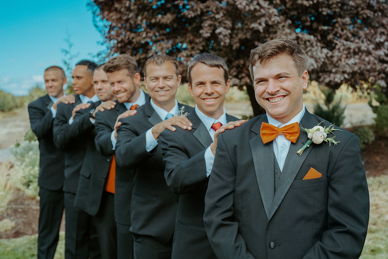 Groom with groomsmen in formal wear, posed outside. They are smiling, with an orange bow tie and pocket square.