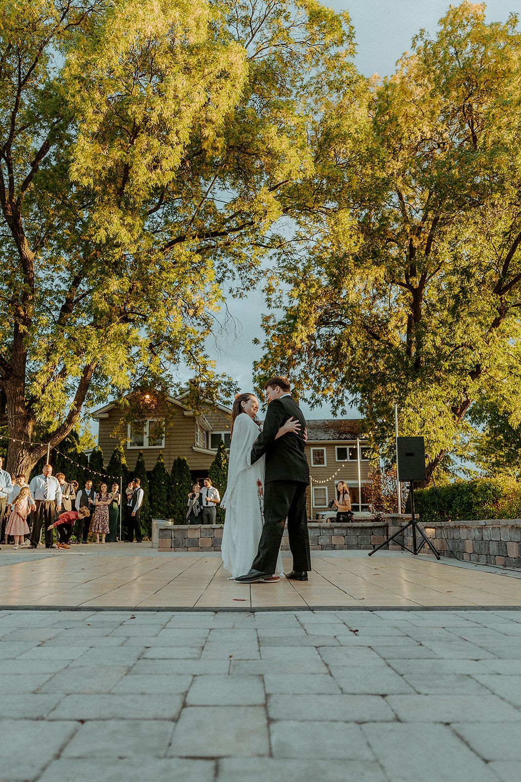 Couple dances on a stone patio, trees framing a house with guests in the background.