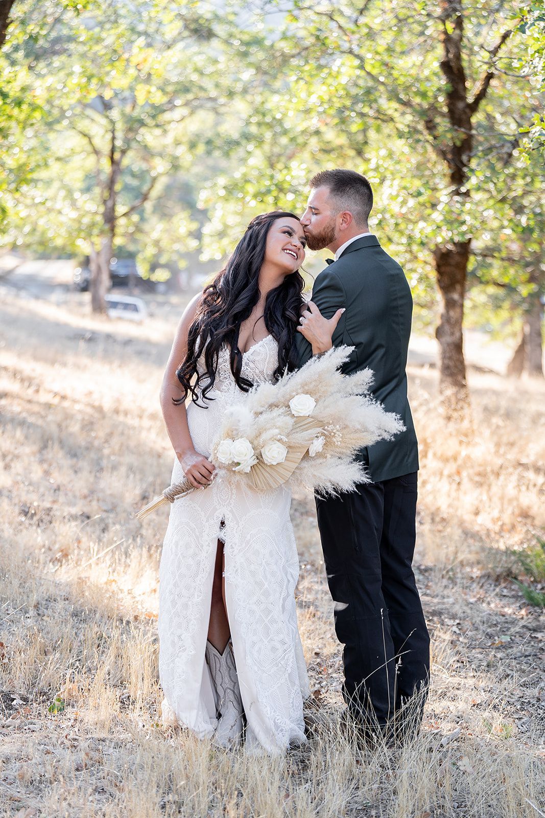 A bride and groom are kissing in a field while the bride is holding a bouquet of pampas grass.