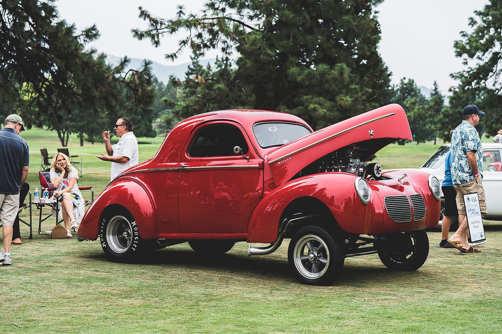 A red car with the hood up is parked in a grassy field.