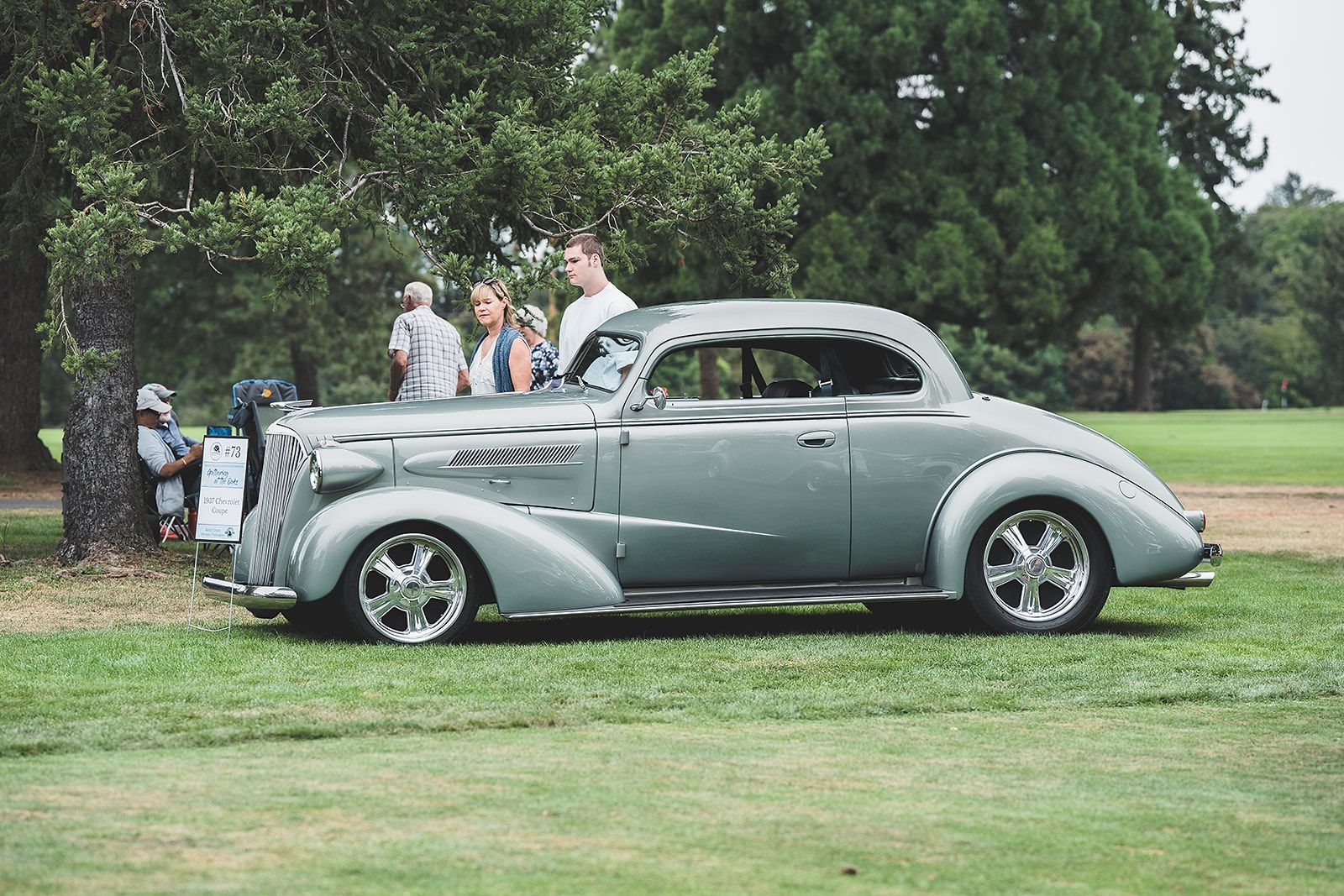 A silver car is parked in a grassy field with people standing around it.