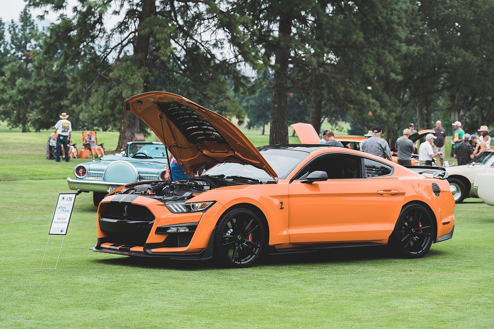An orange mustang with the hood up is parked in a grassy field.