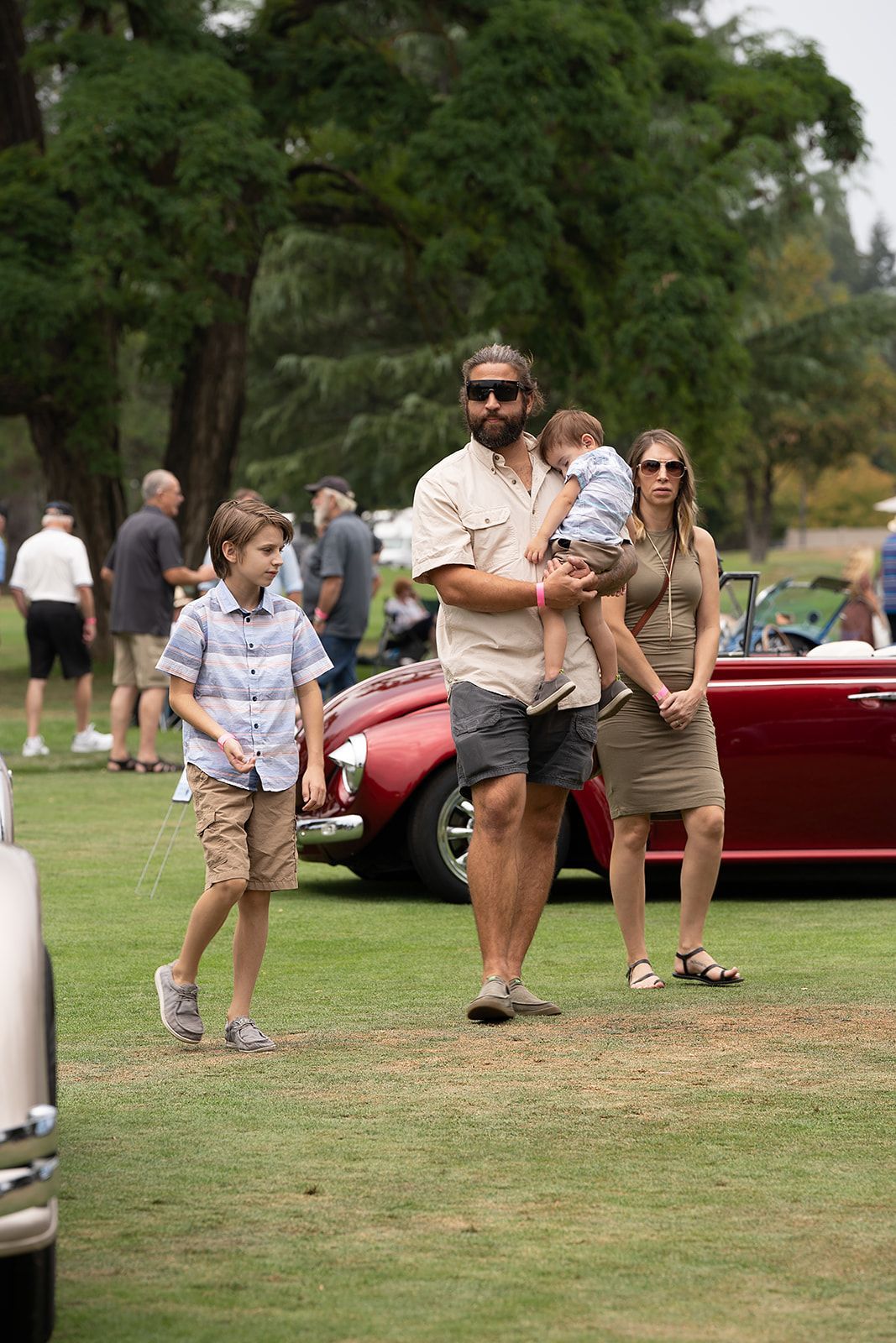 A man is holding a baby while standing next to a woman and two children at a car show.