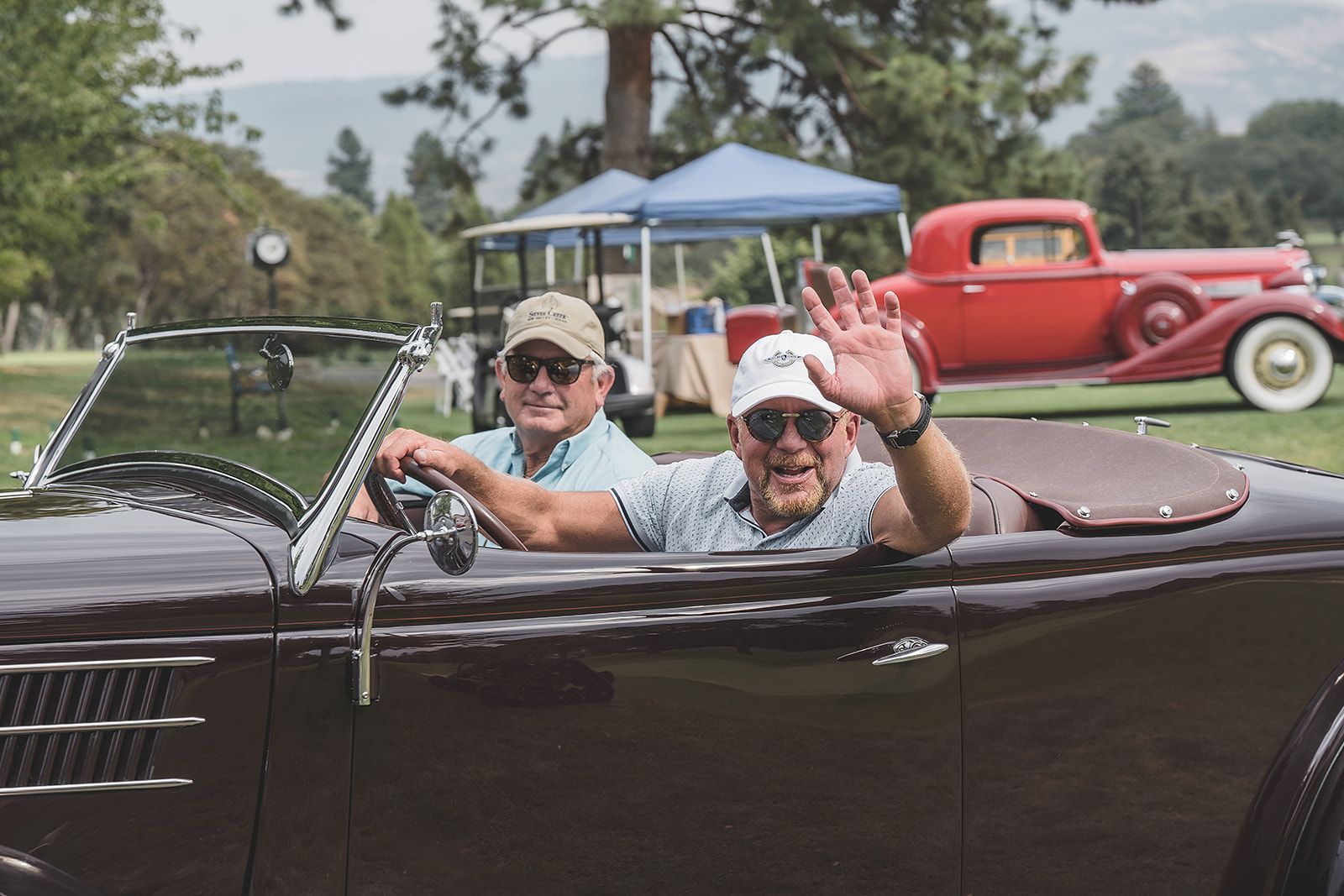 Two men are driving a convertible car and waving from the window.