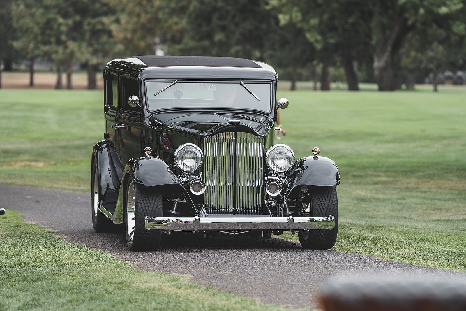 An old black car is driving down a dirt road.
