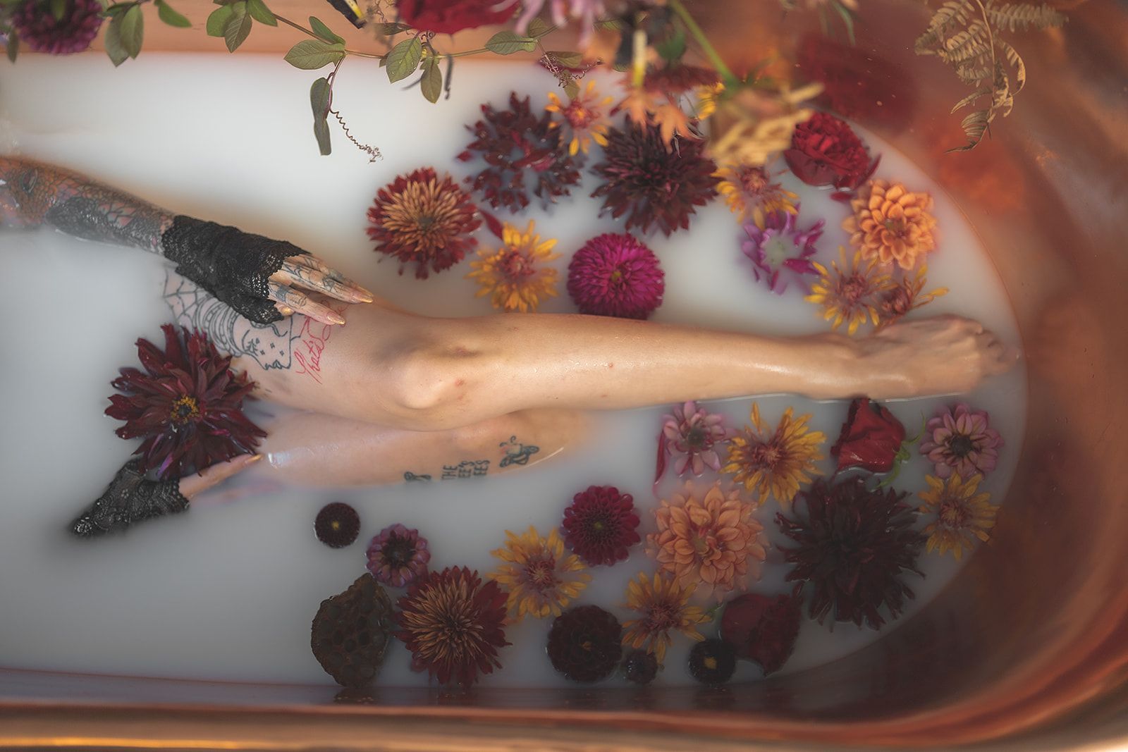 Person in copper tub with flowers in milky water; tattooed arm visible.
