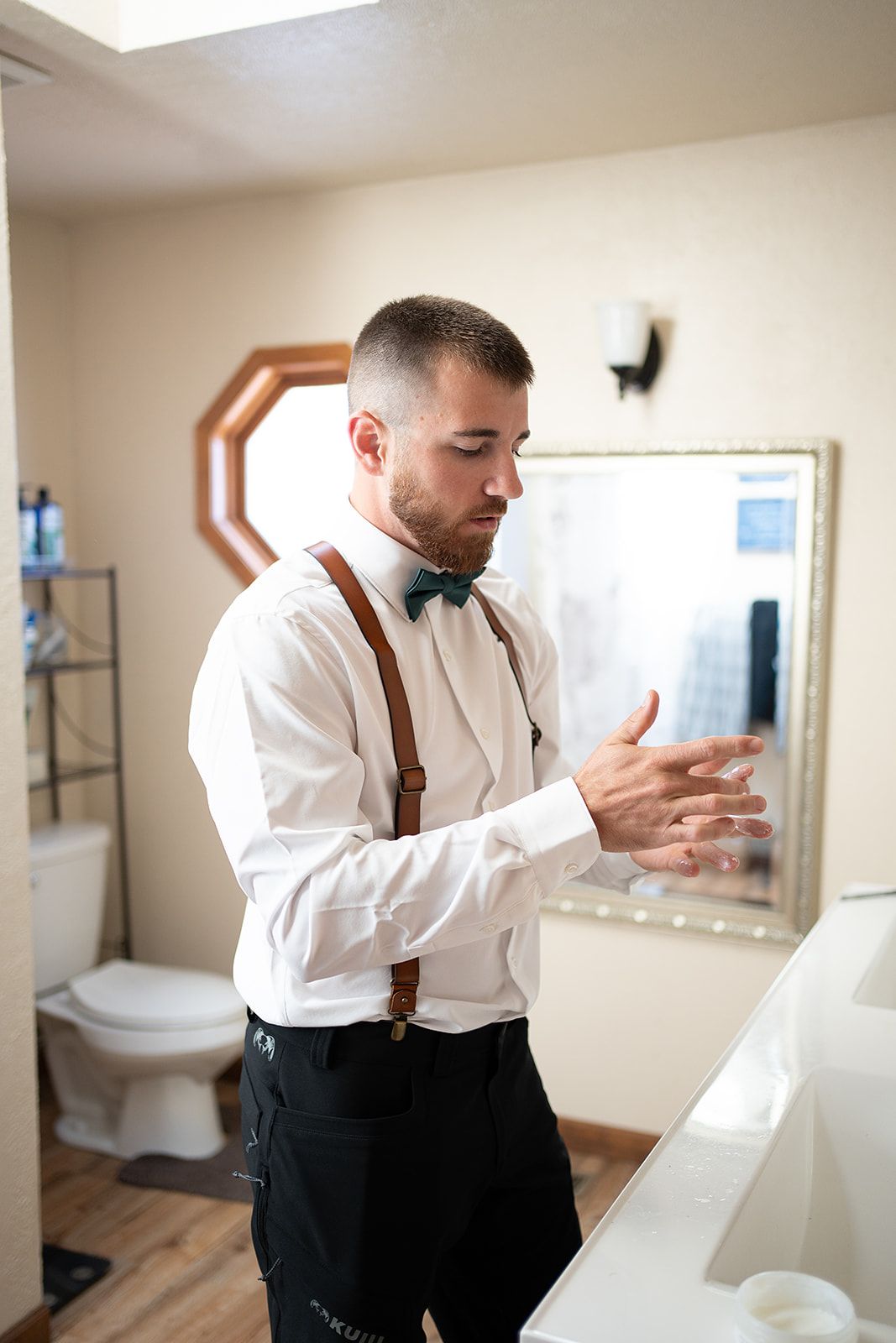 A man in a bow tie and suspenders is standing in a bathroom.