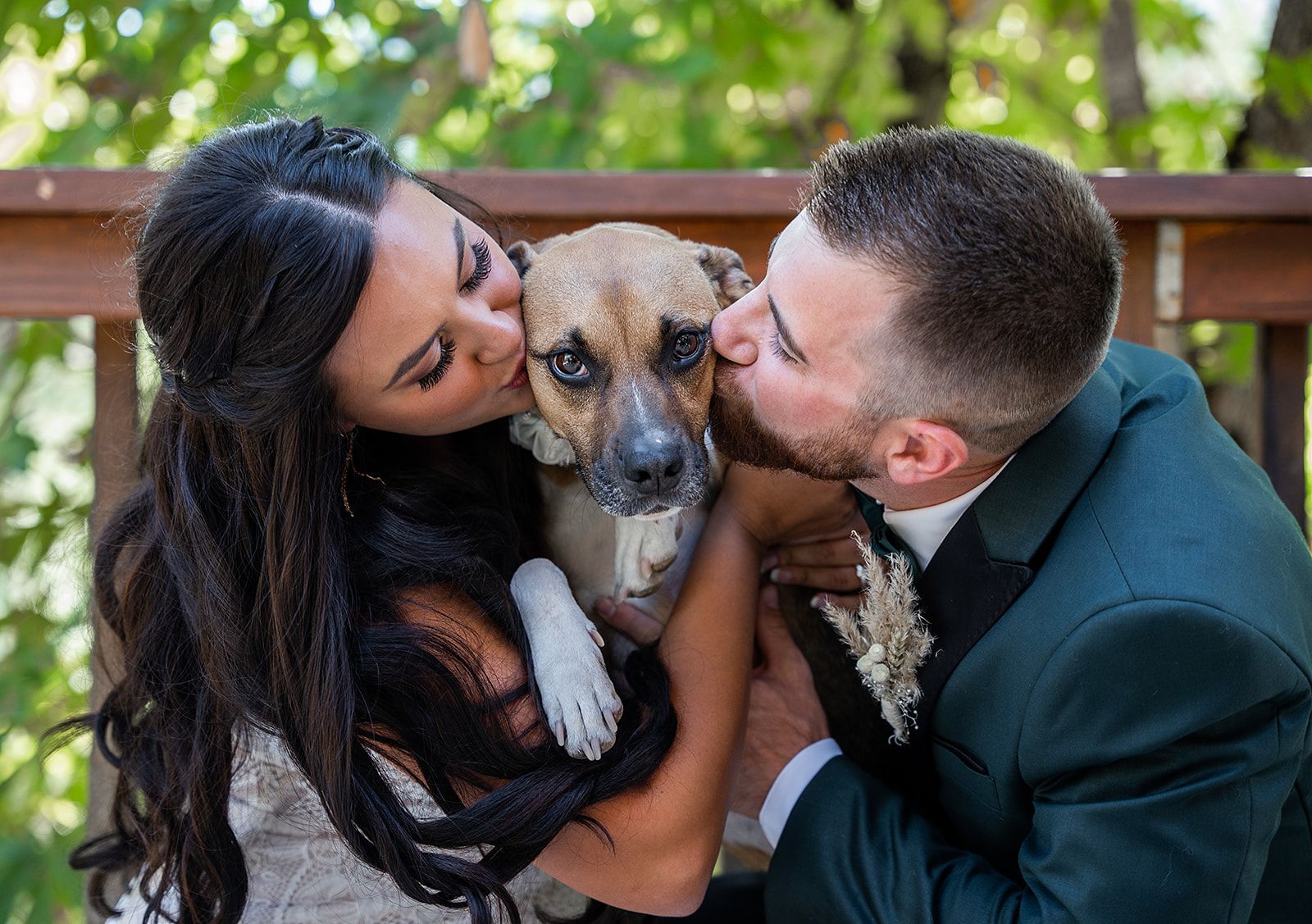 A bride and groom are kissing their dog on the nose.