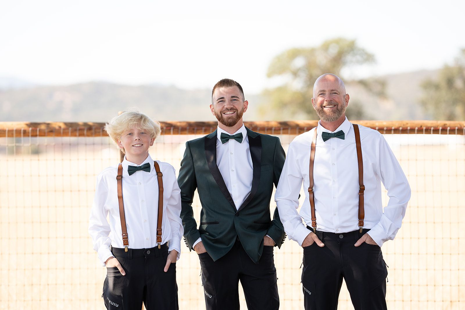 A groom and two groomsmen are standing next to each other wearing suspenders and bow ties.