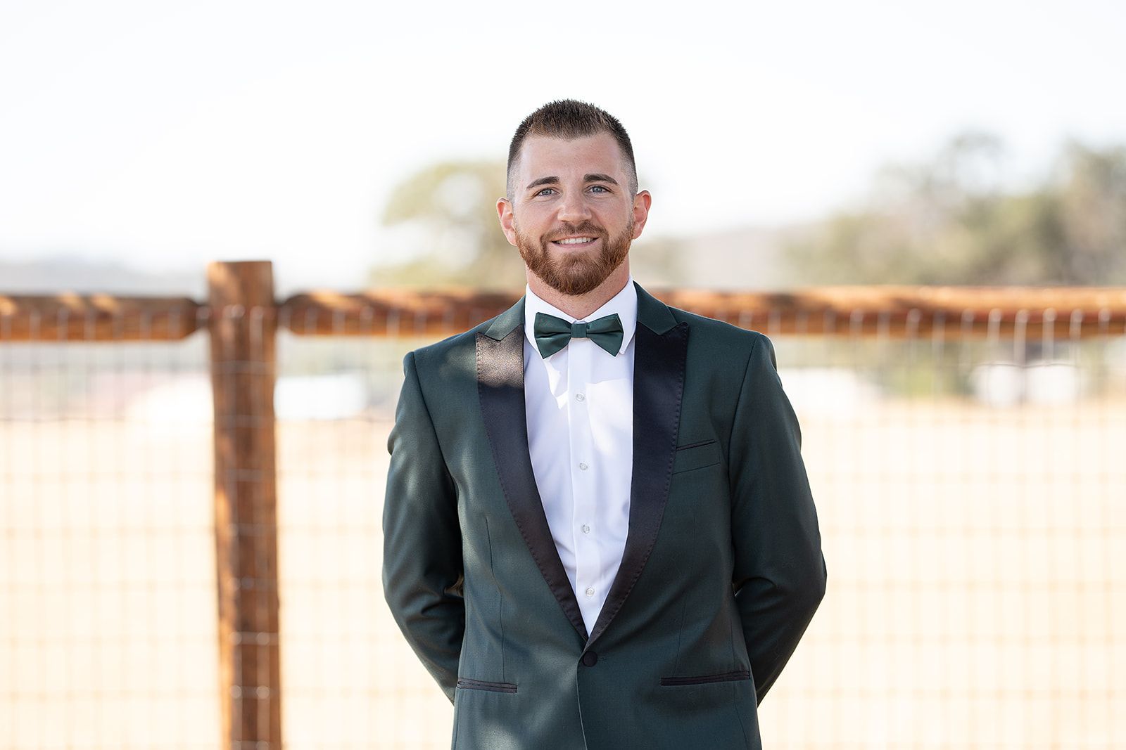 A man in a suit and bow tie is standing in front of a fence.