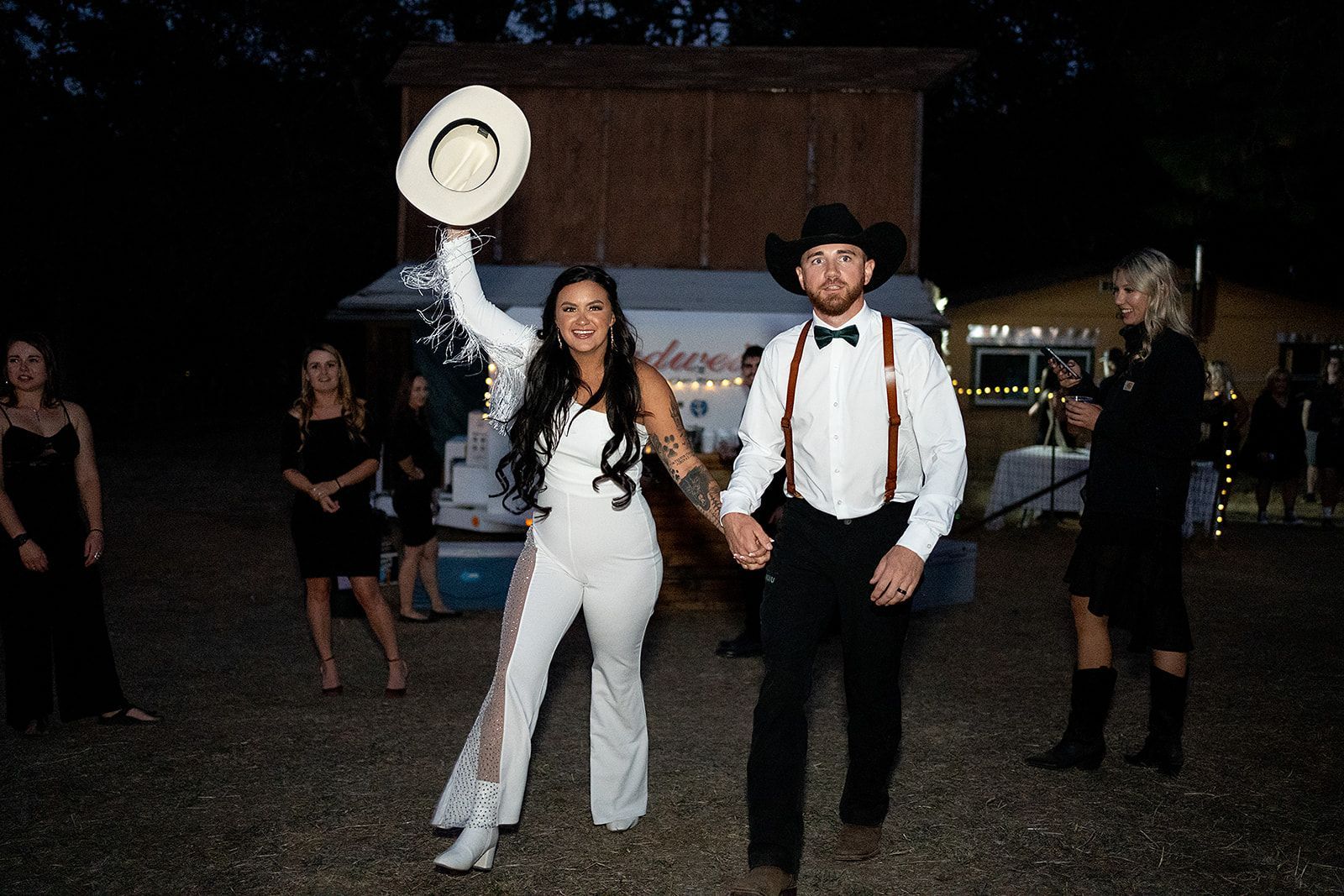 A bride and groom are holding hands and the bride is holding a cowboy hat in the air.