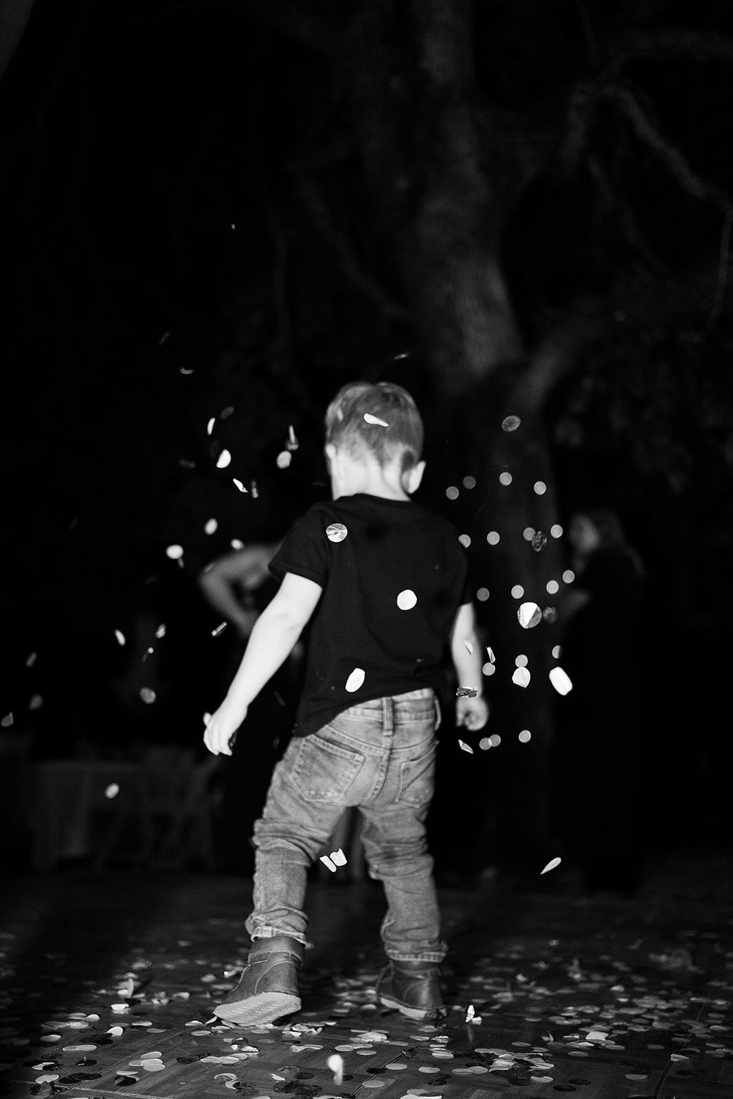A black and white photo of a young boy standing in the dark surrounded by confetti.