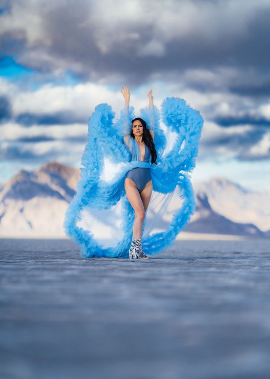 A woman in a blue dress is standing in the middle of a desert holding a camera.