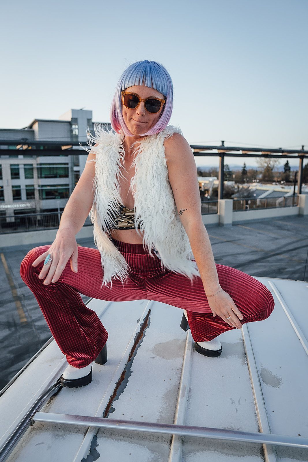 A woman with pink hair and sunglasses is squatting on a rooftop.
