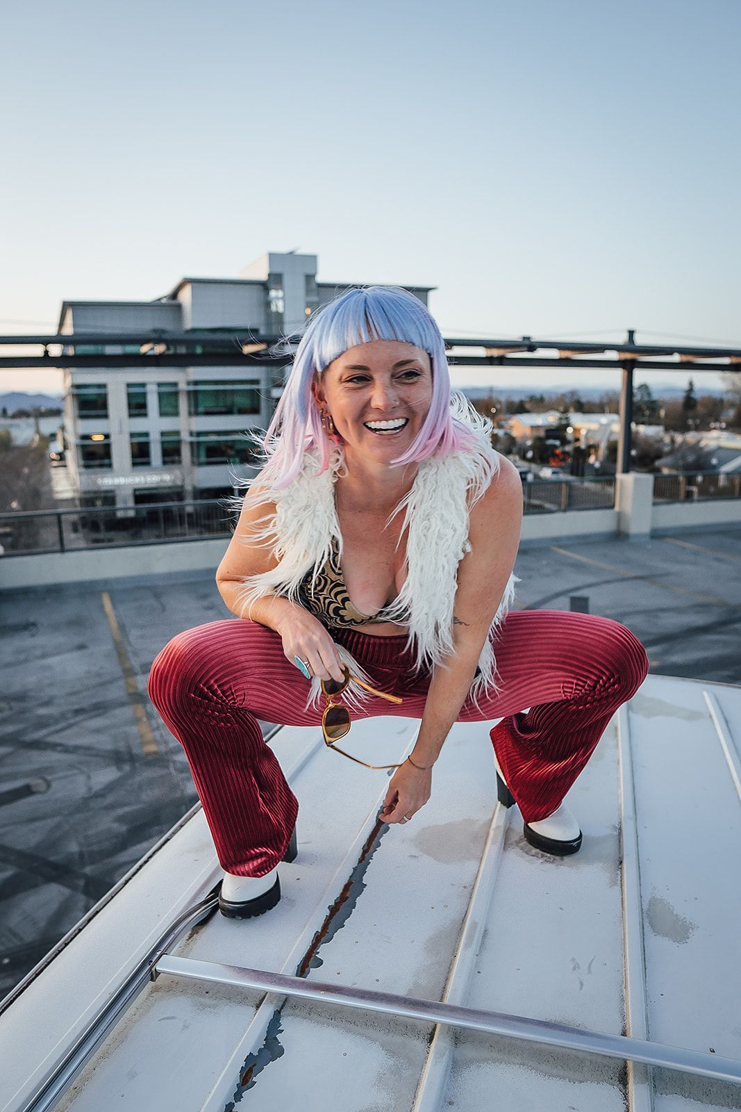 A woman with pink hair and sunglasses is squatting on a rooftop.