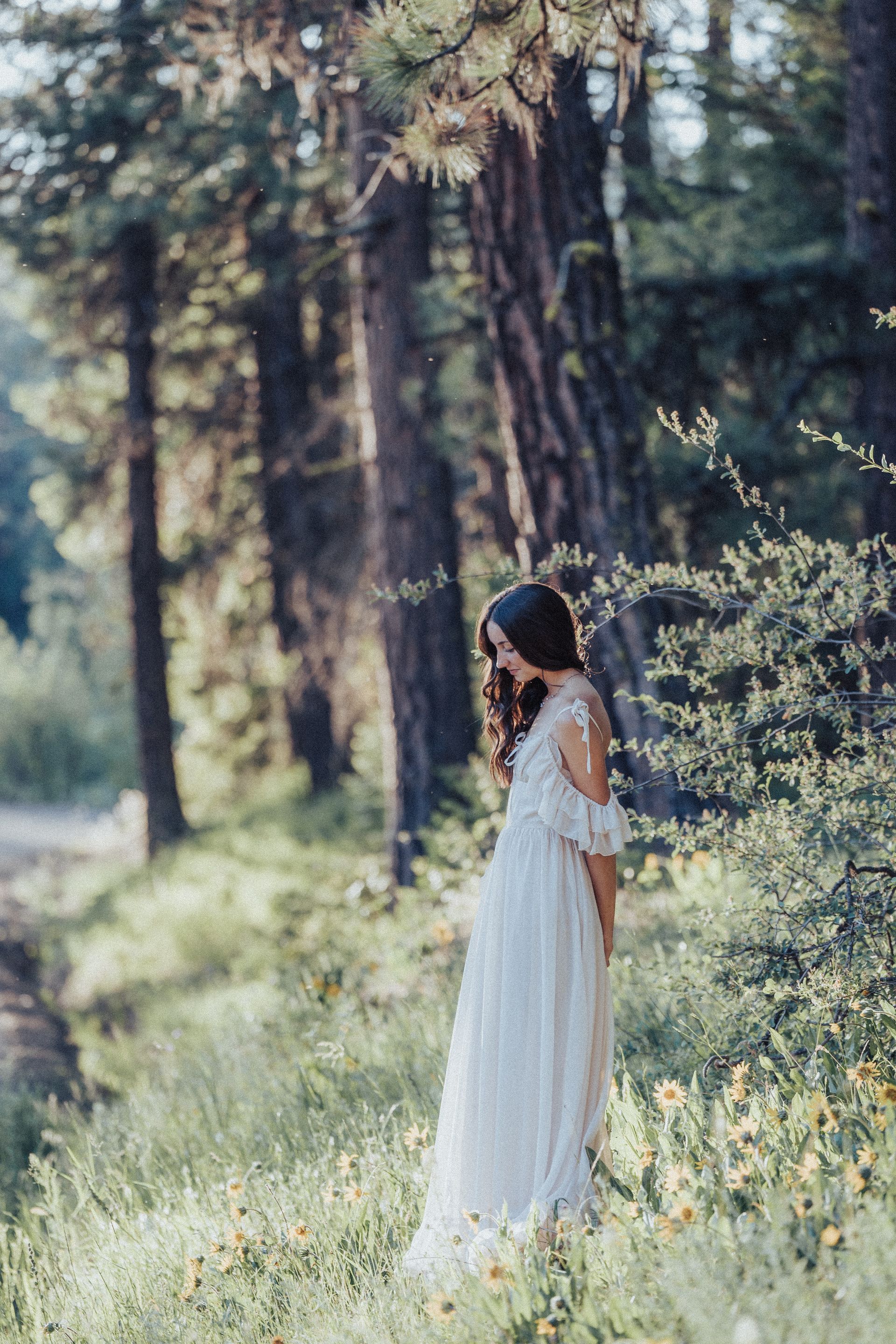 A woman in a white dress is standing in a field of flowers in the woods.