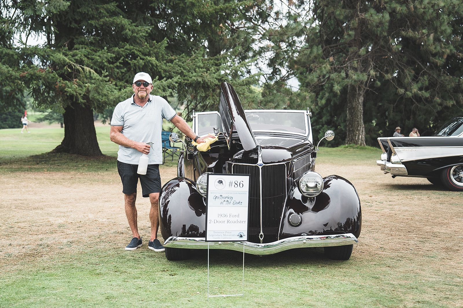 A man is standing in front of a black car with the hood up.