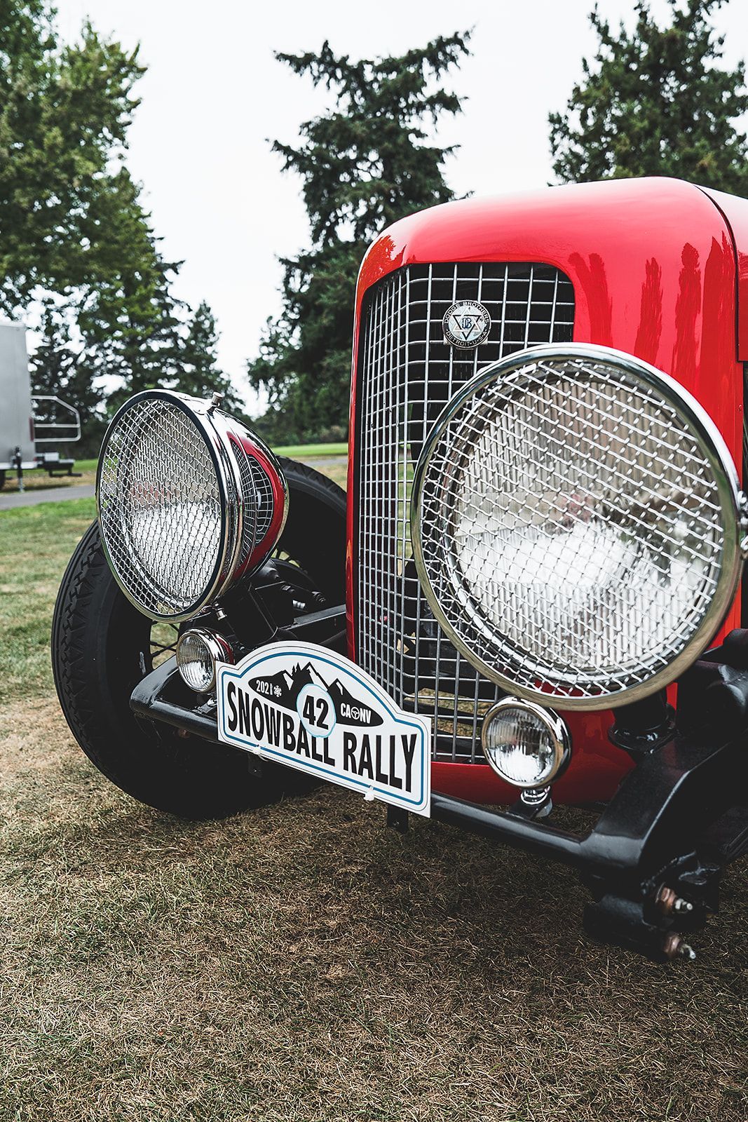 A red car with a license plate that says snowball rally is parked on a gravel road.