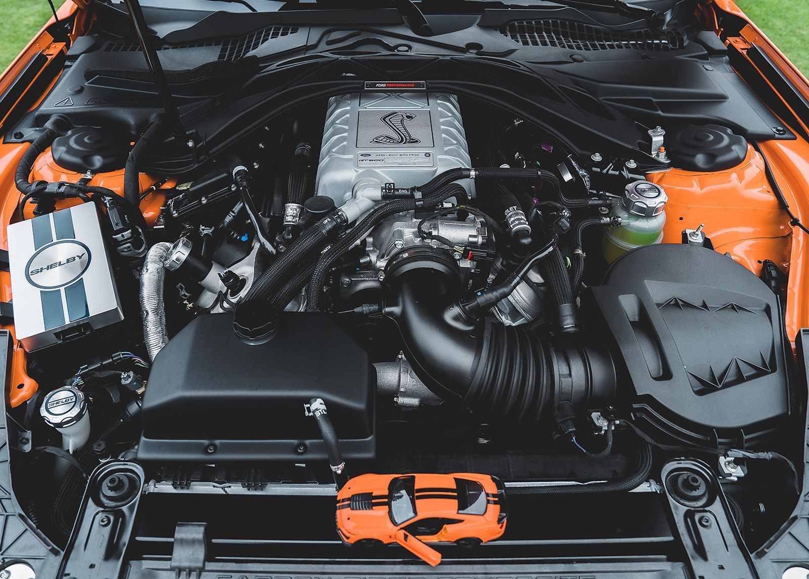 The engine of an orange mustang is shown with the hood open.