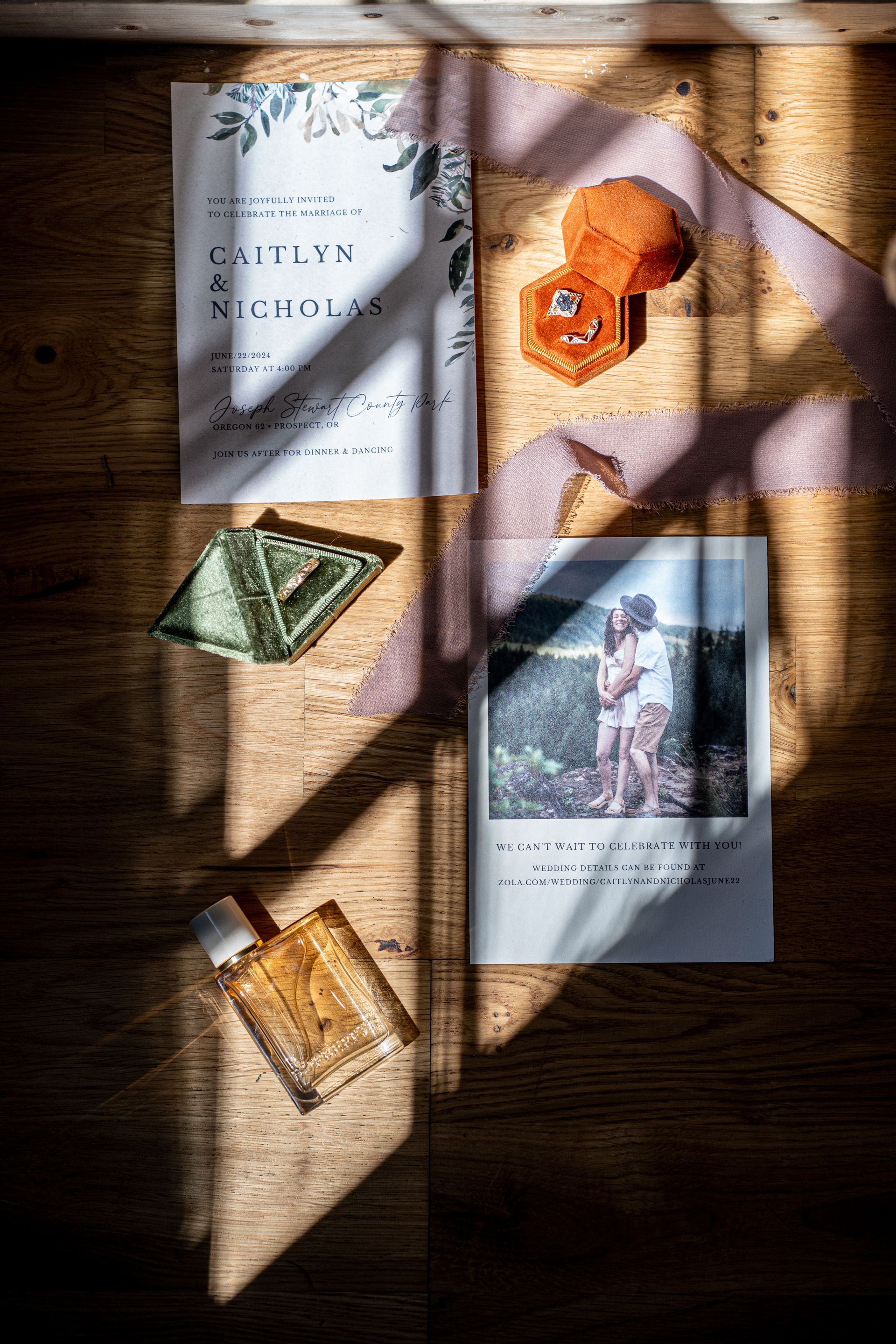 A wedding invitation , rings , perfume , and a picture of a couple are on a wooden table.