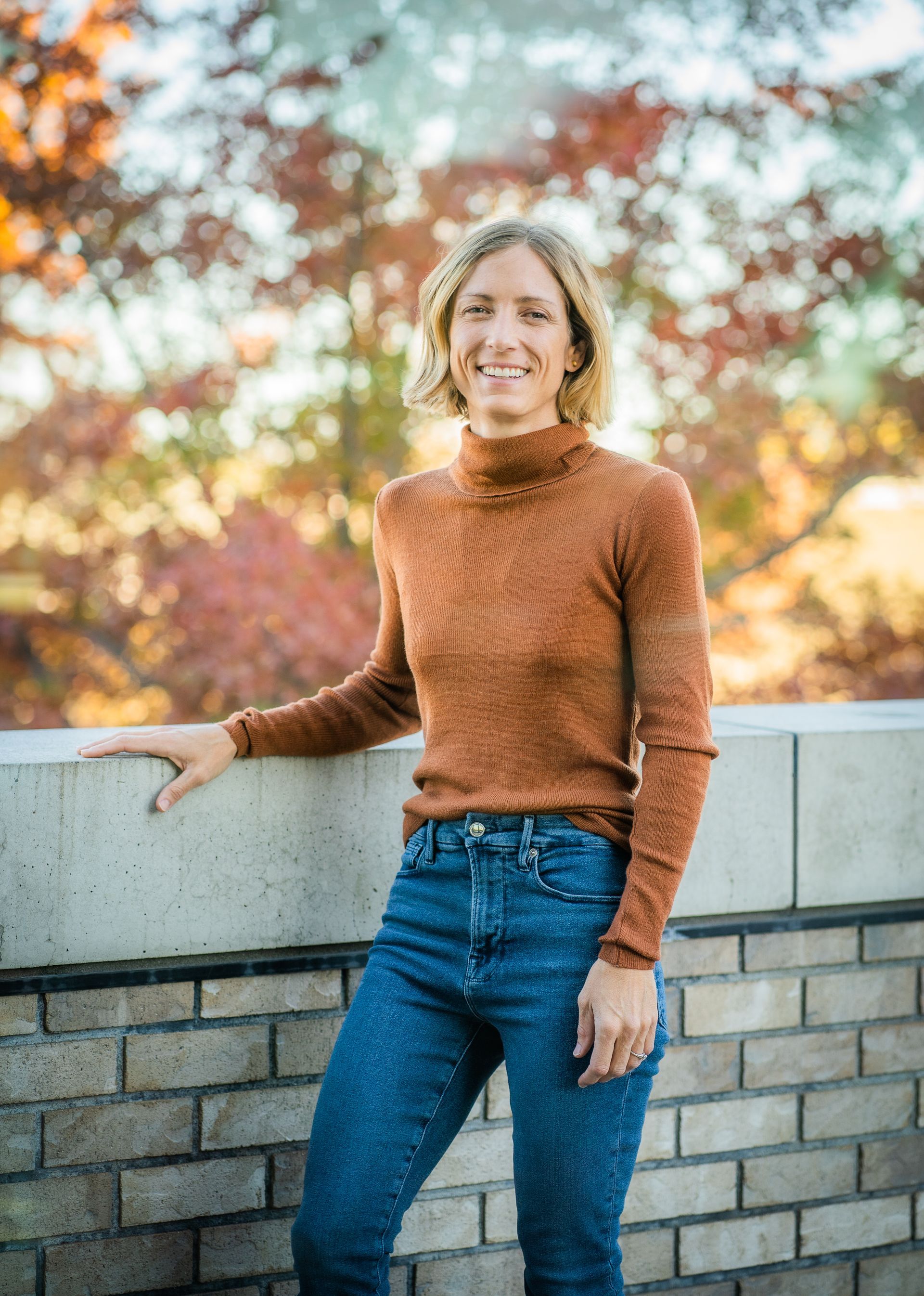 A woman in a brown sweater and blue jeans is standing next to a brick wall.