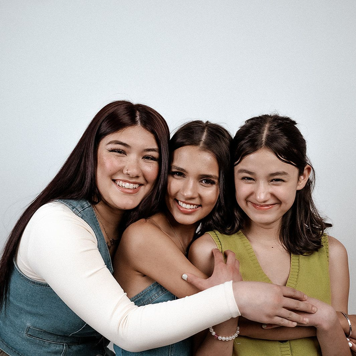 Three smiling people hugging in front of a white backdrop; one in denim, one in blue, one in green.