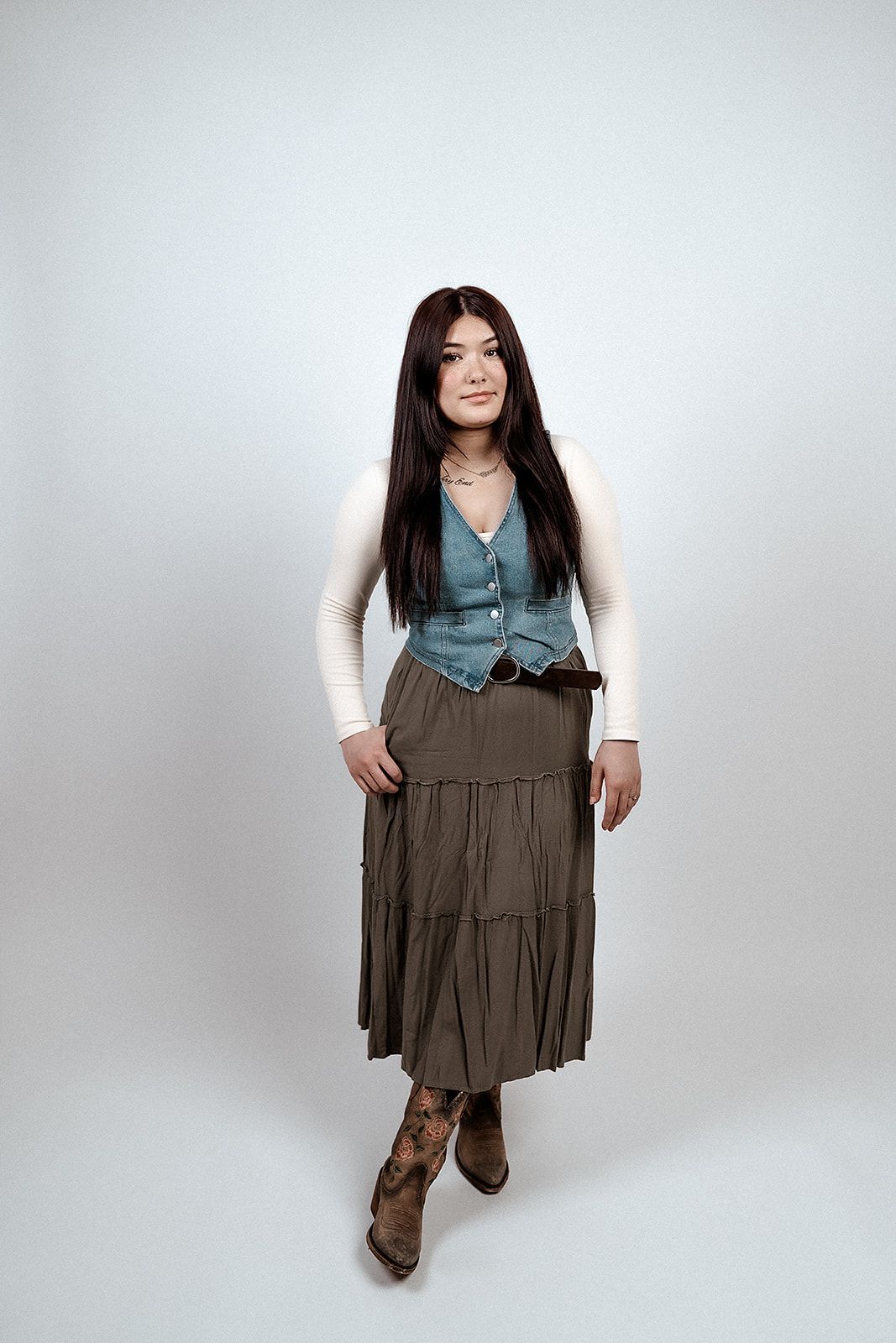 A woman in a skirt and cowboy boots is standing in front of a white background.