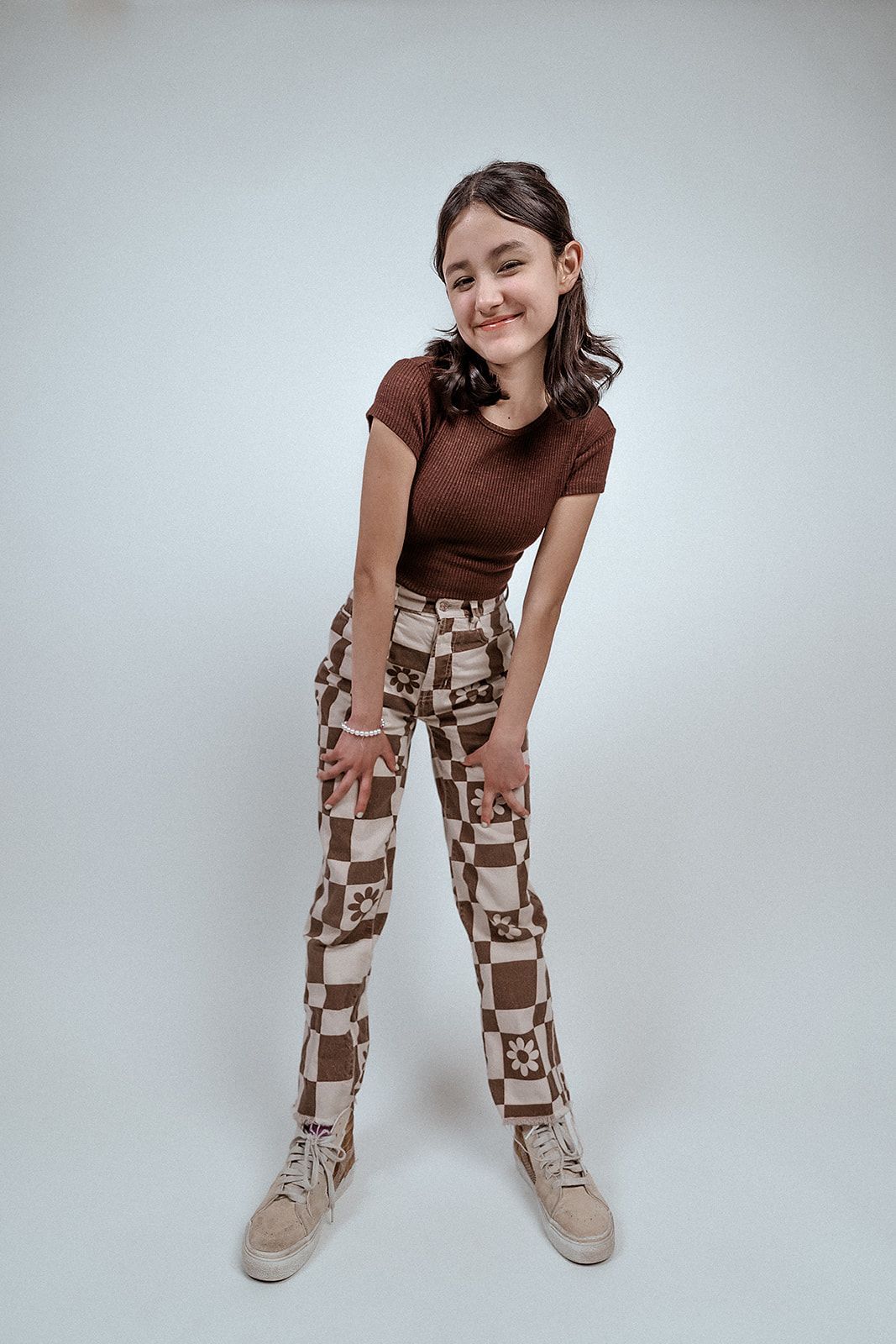 A young woman in a brown top and checkered skirt is standing in front of a white wall.