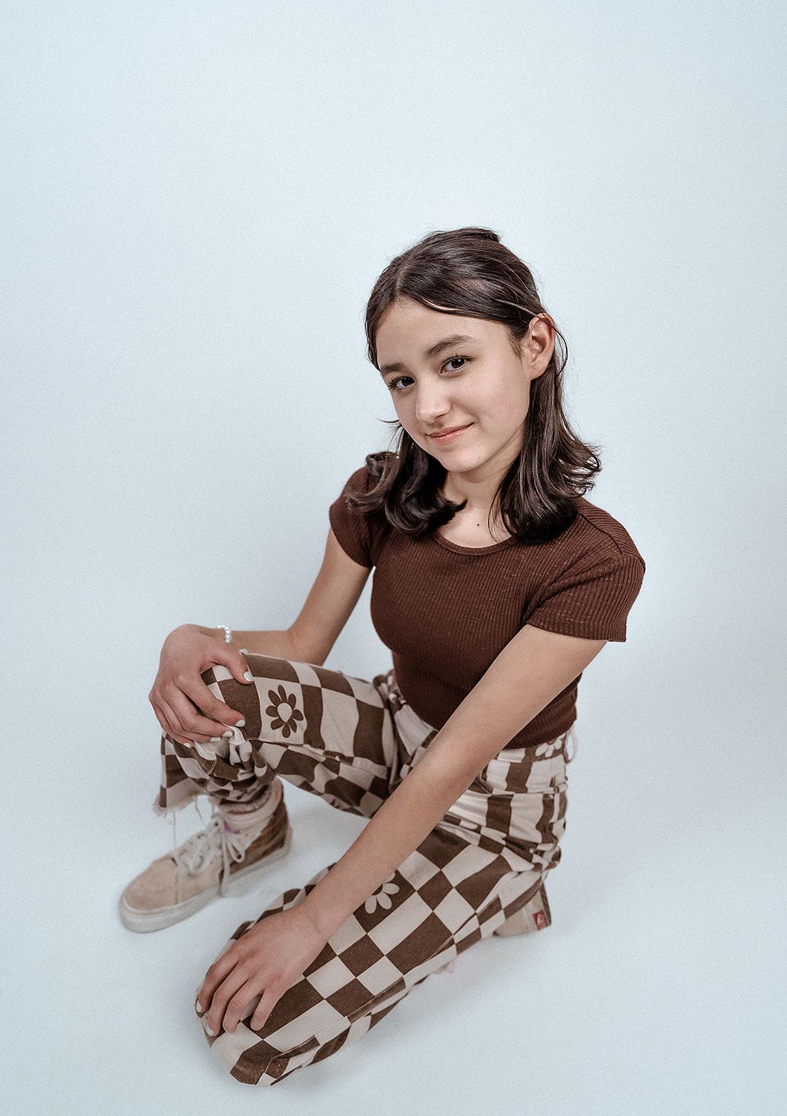 A young girl is sitting on the floor wearing a brown shirt and checkered pants.