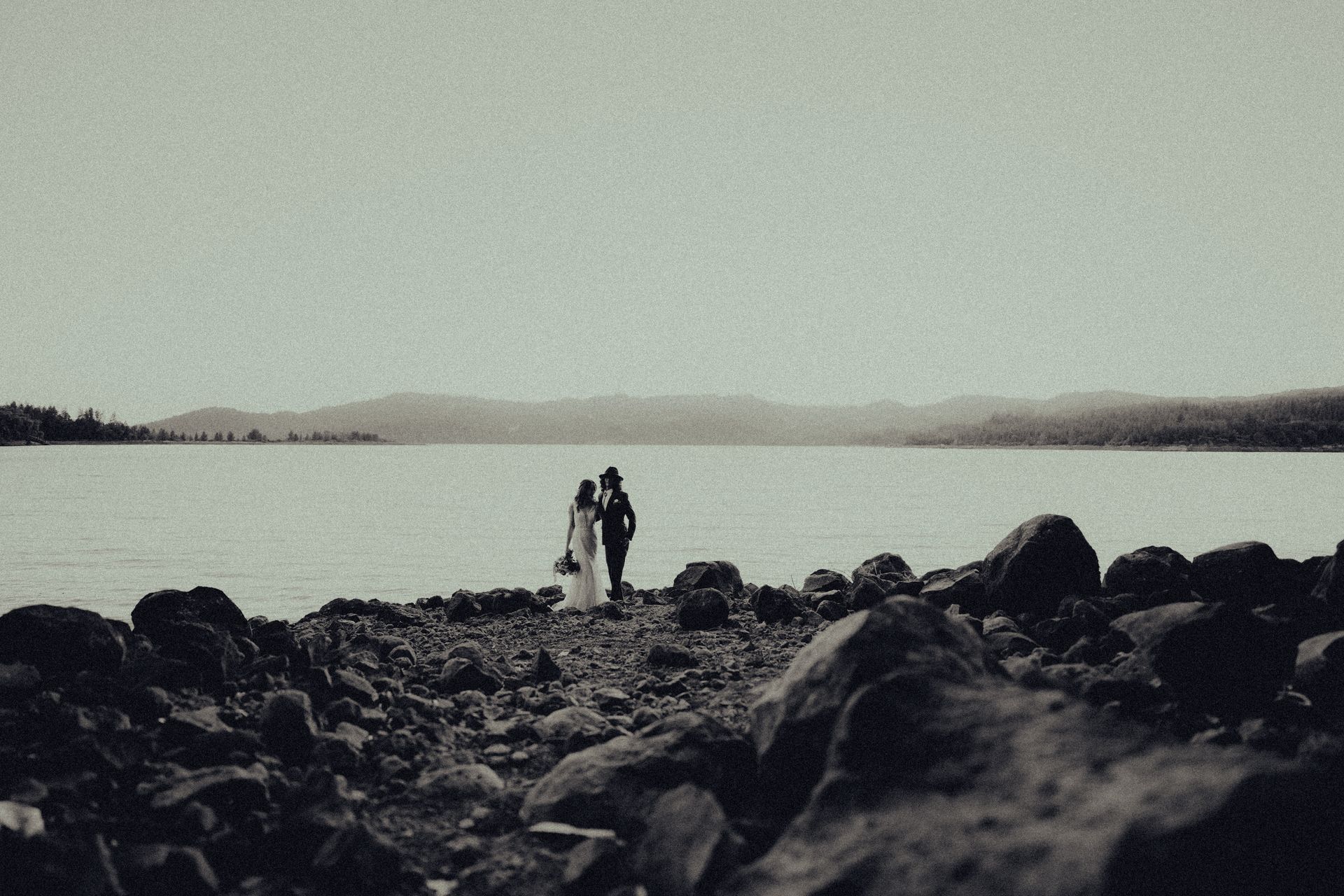A black and white photo of a bride and groom standing on the shore of a lake.