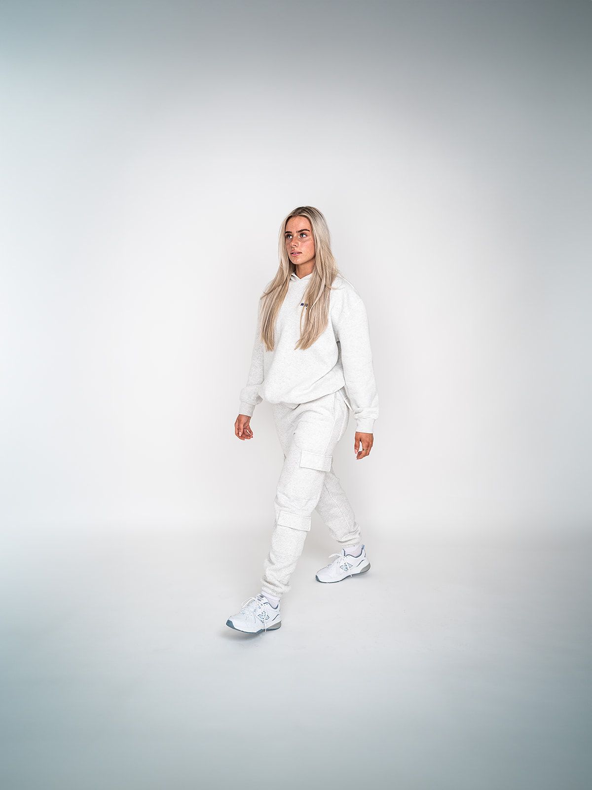 A woman in a white sweatshirt and white sweatpants is standing in front of a white wall.
