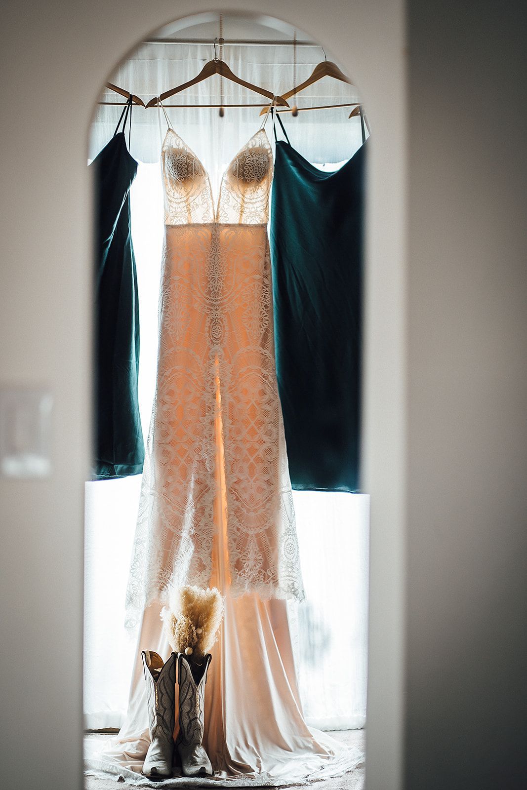 A wedding dress is hanging in a window next to a pair of cowboy boots.