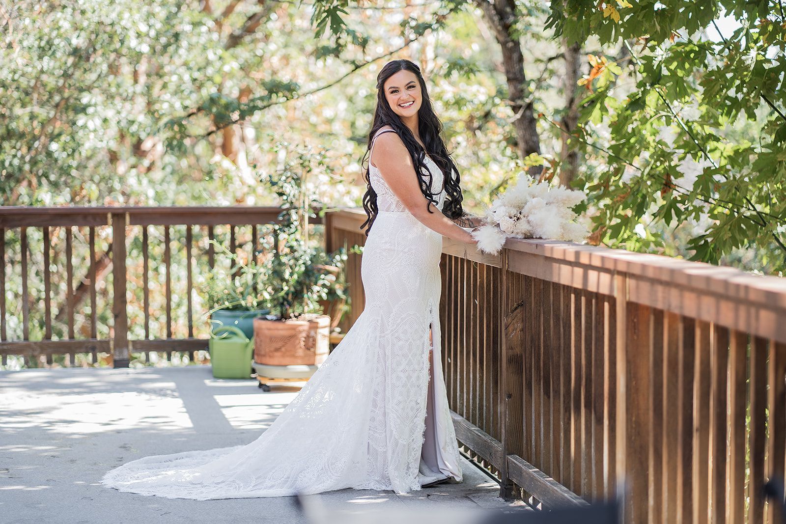 A bride in a wedding dress is leaning against a wooden railing on a deck.