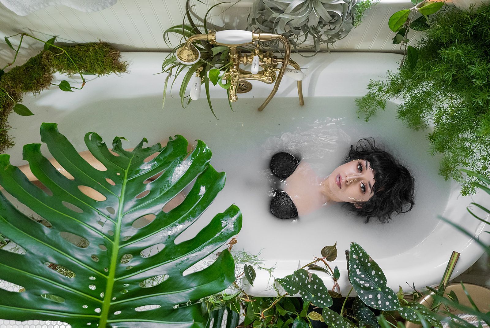 A woman is laying in a bathtub filled with milk, surrounded by plants.
